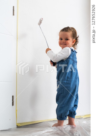 Cute little curly haired girl painting a white wall with a roller, wearing denim overalls and a white sweater. 127652926