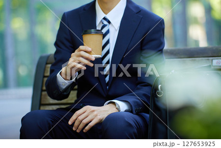 ビジネス街でベンチに座りコーヒーを飲むスーツの若い男性 ビジネス街でベンチに座りコーヒーを飲むスーツの若い男性 127653925
