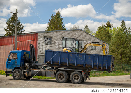 Blue industrial truck carrying a yellow mini excavator in its container bed 127654595