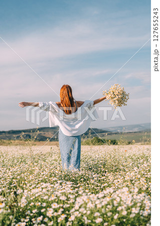Daisies Woman Meadow: Redhead spreads arms holding flowers in daisy field during daytime. Daisies Woman Meadow: Redhead spreads arms holding flowers in daisy field during daytime. 127655243
