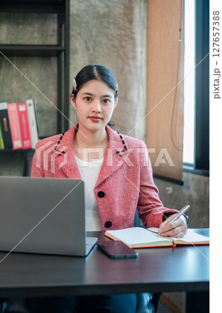 Businesswoman in a pink blazer working at a desk with a laptop and notebook in a contemporary office environment. Businesswoman in a pink blazer working at a desk with a laptop and notebook in a contemporary office environment. 127657388