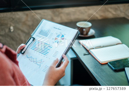 Close-up of a person reviewing financial charts and graphs, with a notebook and coffee on a desk. 127657389