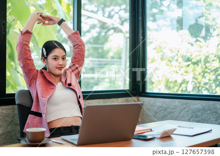 A young woman in a pink blazer stretches at her desk in a bright, modern office setting with a laptop and coffee. A young woman in a pink blazer stretches at her desk in a bright, modern office setting with a laptop and coffee. 127657398