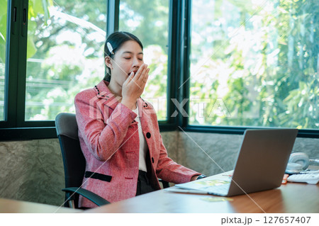 A young professional woman yawning at her desk in a modern office setting, surrounded by large windows and greenery. 127657407