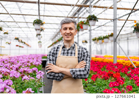A smiling man in an apron stands in a greenhouse, surrounded by colorful flowers. A smiling man in an apron stands in a greenhouse, surrounded by colorful flowers. 127661041