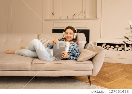 Teenage girl lounging on sofa wearing headphones holding tablet device. 127661438