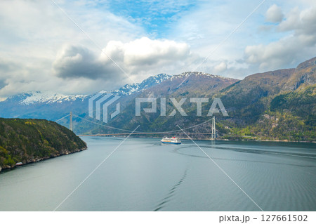 Distant tourist ship sails under Hardanger Bridge gracefully spanning waters of fjord. Mountains with snow on tops create Norwegian landscape Distant tourist ship sails under Hardanger Bridge gracefully spanning waters of fjord. Mountains with snow on tops create Norwegian landscape 127661502