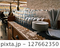 Close-up of clean glasses, utensils, stacked plates on weathered wooden countertop at outdoor cafe, Lisbon, Portugal. Relaxed dining atmosphere, people seated, umbrellas, historic city square, travel 127662350