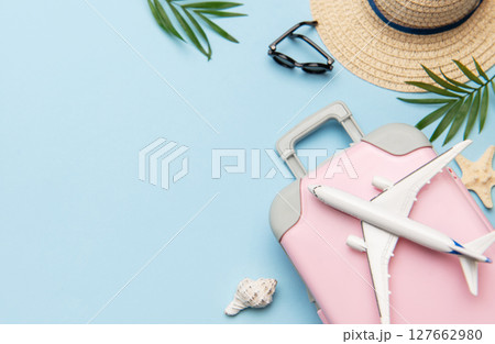 Pink suitcase with toy airplane, sunglasses, straw hat, seashells and palm leaves on blue background Pink suitcase with toy airplane, sunglasses, straw hat, seashells and palm leaves on blue background 127662980