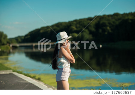 A slender girl by the water, dressed in a T-shirt and shorts, holds a phone and talks 127663657
