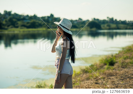A young lady in white clothes stands against the backdrop of a lake, having a lively phone conversation 127663659