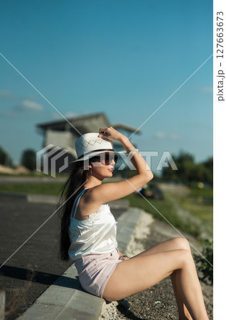 The slender girl is sitting on the warm soil, shielding her eyes from the sun with stylish glasses 127663673