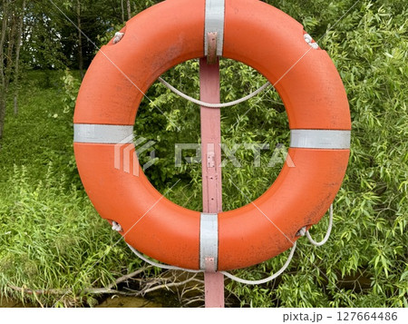 An orange and white lifebuoy hangs on the dock against a backdrop of blooming greenery by the river. High quality photo An orange and white lifebuoy hangs on the dock against a backdrop of blooming greenery by the river. High quality photo 127664486