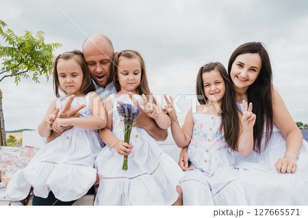 The father, mother, and their three daughters sit on a wooden bench in a blooming field The father, mother, and their three daughters sit on a wooden bench in a blooming field 127665571