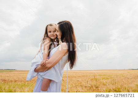 A young mother in a long white dress holds her little daughter in her arms in a field of barley A young mother in a long white dress holds her little daughter in her arms in a field of barley 127665721