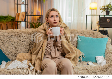 Woman covered with blanket on sofa, warming hands on tea cup, showing cold symptoms with tired look 127665882