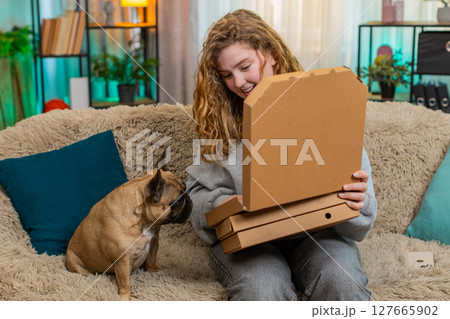 Young woman opening pizza boxes on home sofa while pug dog bites slice with playful hungry joy Young woman opening pizza boxes on home sofa while pug dog bites slice with playful hungry joy 127665902