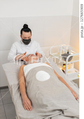 Woman completing facial wrap mask procedure in cosmetology clinic, beautician apply gauze skin care Woman completing facial wrap mask procedure in cosmetology clinic, beautician apply gauze skin care 127665909