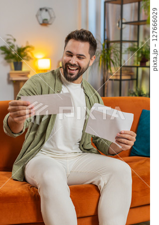 Excited young man opening envelope letter and reading good news sitting on sofa at home apartment Excited young man opening envelope letter and reading good news sitting on sofa at home apartment 127666029