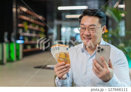 Successful businessman holding smartphone and counting euro bill money fan sitting in office lobby Successful businessman holding smartphone and counting euro bill money fan sitting in office lobby 127666111