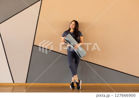Curvy woman in activewear leaning on wall with yoga mat, smiling before workout 127666900