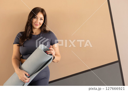 Full-figured woman in gym clothes posing with yoga mat near wall after training session Full-figured woman in gym clothes posing with yoga mat near wall after training session 127666921