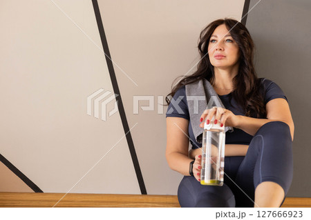 Fit woman in sportswear resting after workout with water bottle, sitting and smiling in gym interior 127666923