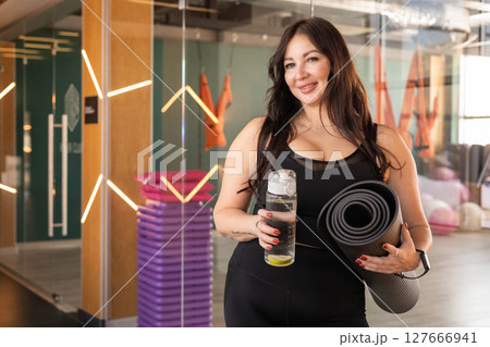 Fit woman holding yoga mat and water bottle after class in modern wellness and fitness studio Fit woman holding yoga mat and water bottle after class in modern wellness and fitness studio 127666941