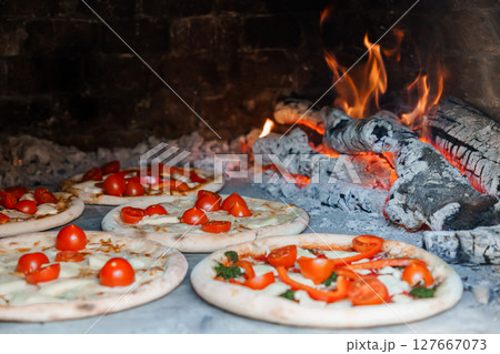 Wood-Fired Pizza Baking in Brick Oven with Flames. Rustic Italian Cuisine, Fresh Tomatoes, Molten Cheese, Close-Up Food Photography 127667073