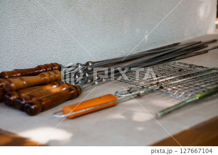 Rustic BBQ Tools Laid Out on Stone Countertop. Grills, Skewers, Tongs, Wood Handles, Metal Texture, Brown and Gray Culinary Still Life. Rustic BBQ Tools Laid Out on Stone Countertop. Grills, Skewers, Tongs, Wood Handles, Metal Texture, Brown and Gray Culinary Still Life. 127667104