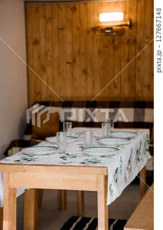 Rustic Wooden Dining Scene: Family Meal Setup with Checkered Bench, Light Wood Table, Plates and Greenery in Cozy Cabin Interior. 127667148