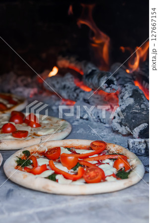Rustic Wood-Fired Pizza with Fresh Tomatoes and Basil: Close-Up of Artisanal Pies Baking in Stone Oven with Burning Embers. 127667154