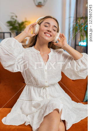 Joyful young Caucasian woman listening to music through wireless headphones sitting on sofa couch Joyful young Caucasian woman listening to music through wireless headphones sitting on sofa couch 127667245