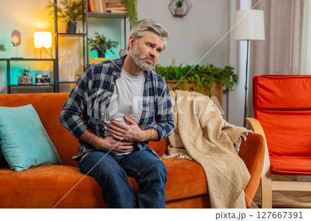 Senior middle-aged man sits on couch feeling sudden strong abdominal stomach ache gastritis problem 127667391