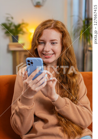 Happy young woman sitting on sofa using smartphone texting smiling at home living room apartment 127667415