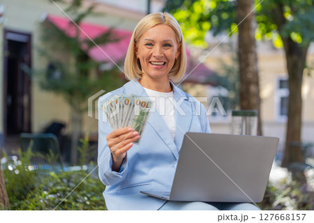 Happy mature Caucasian businesswoman CEO holding cash and using laptop computer on city street 127668157