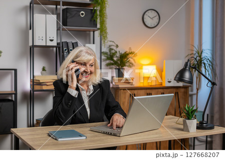 Senior Caucasian businesswoman operator using smartphone and laptop computer at home office desk Senior Caucasian businesswoman operator using smartphone and laptop computer at home office desk 127668207