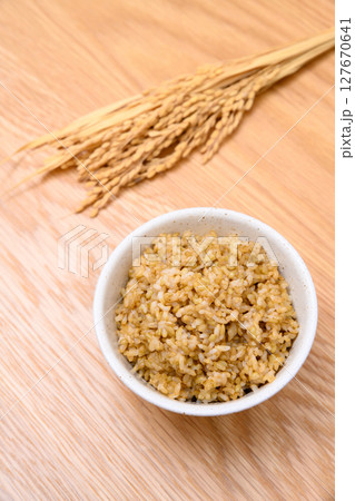 木目テーブル背景の茶碗に玄米ごはんと稲穂 127670641
