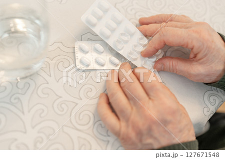 Senior person hands holding blister packs of pills over a patterned tablecloth, with a glass of water nearby, copy space. Concept of senior health care and daily medication routine Senior person hands holding blister packs of pills over a patterned tablecloth, with a glass of water nearby, copy space. Concept of senior health care and daily medication routine 127671548