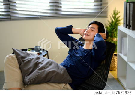 Young man relaxing in a chair with headphones on, enjoying music in a cozy home Young man relaxing in a chair with headphones on, enjoying music in a cozy home 127671972