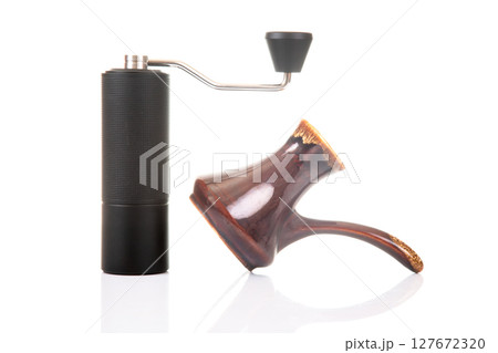 A black manual coffee grinder with a metallic handle stands next to a copper cezve A black manual coffee grinder with a metallic handle stands next to a copper cezve 127672320