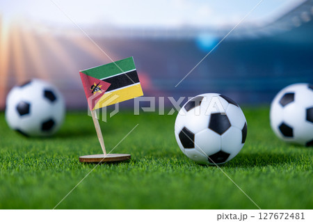 Miniature Mozambique flag with multiple soccer balls on green grass turf, symbolizing national team participation and global football competition within a stadium arena 127672481