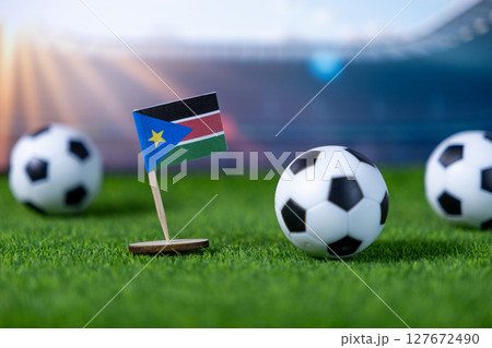 South Sudan flag miniature on artificial green turf with small soccer balls, representing national football team participation in stadium competition South Sudan flag miniature on artificial green turf with small soccer balls, representing national football team participation in stadium competition 127672490