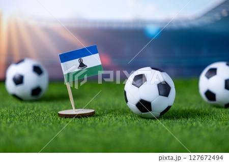 Lesotho national flag miniature prominently placed on vibrant green artificial grass with classic soccer balls, representing global football competition 127672494