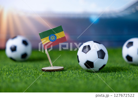Small Ethiopian flag standing proudly on green grass with miniature soccer balls at a blurred stadium, symbolizing national team sports pride and international football events Small Ethiopian flag standing proudly on green grass with miniature soccer balls at a blurred stadium, symbolizing national team sports pride and international football events 127672522