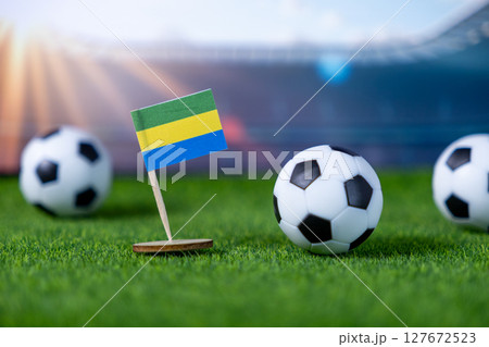 Small Gabon flag stands with mini soccer balls on green artificial grass, symbolizing global football competition and national team pride at a stadium venue 127672523