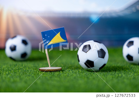 Small Tokelau flag stands firmly on green artificial grass with soccer balls, representing global sports competition and national team spirit in a stadium 127672533