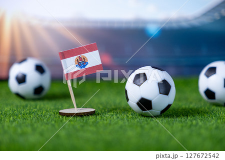 French Polynesia flag miniature stands on green grass with soccer balls and stadium in background, symbolizing the nation's team in global sports 127672542