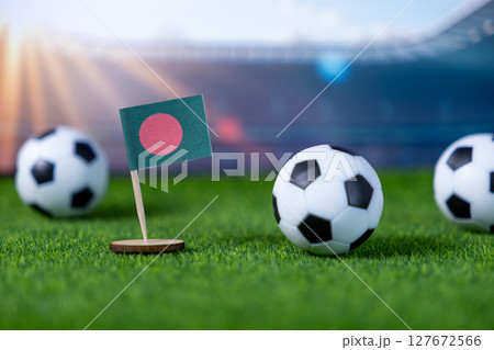 Vibrant miniature Bangladesh flag stands on green artificial turf amidst small soccer balls, symbolizing national team pride, international sport, and global football competition 127672566
