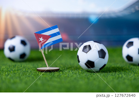 A Cuban flag stands firmly on green artificial grass with miniature soccer balls, portraying national representation and excitement for international football events in a stadium setting 127672640